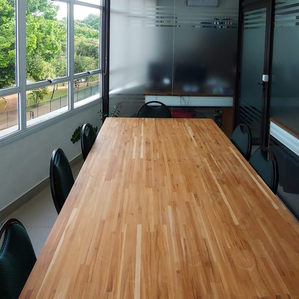 Interior de uma sala de reunião no Trampô Space Coworking, com foco na longa mesa de madeira que convida à colaboração, flanqueada por cadeiras ergonômicas e complementada pela luz natural e pela vista calmante da folhagem externa através da janela