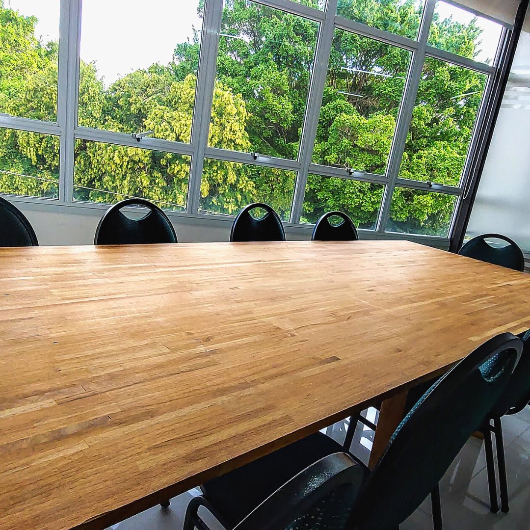 Mesa de reunião de madeira posicionada em uma sala com amplas janelas que revelam uma exuberante paisagem verde, proporcionando um retiro tranquilo e natural para deliberações e estratégias de negócios no interior do Trampô Space Coworking.
